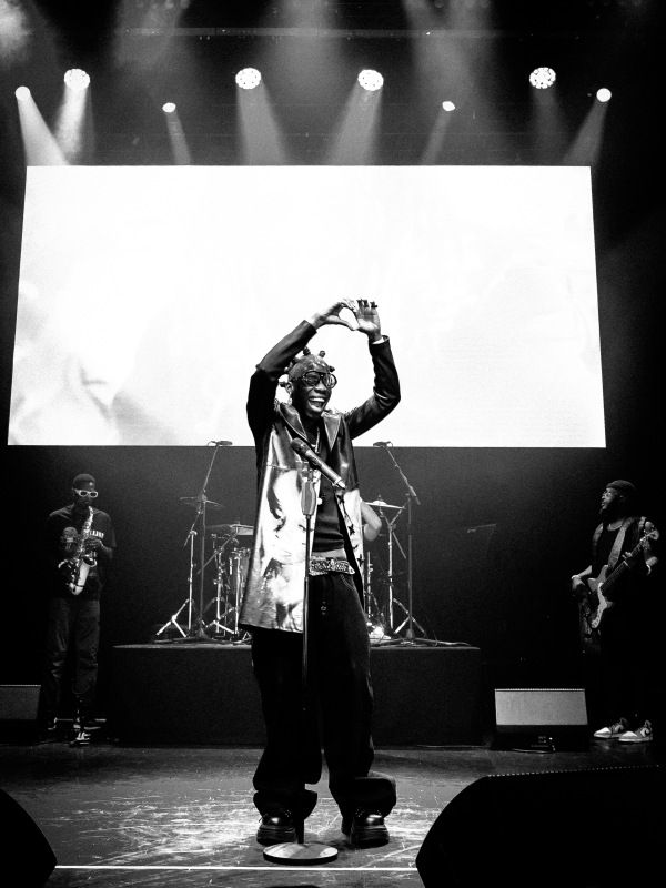 Black and white wide shot of a performer on stage making a heart gesture with their hands for the audience. — concert photography London by Lamss Media